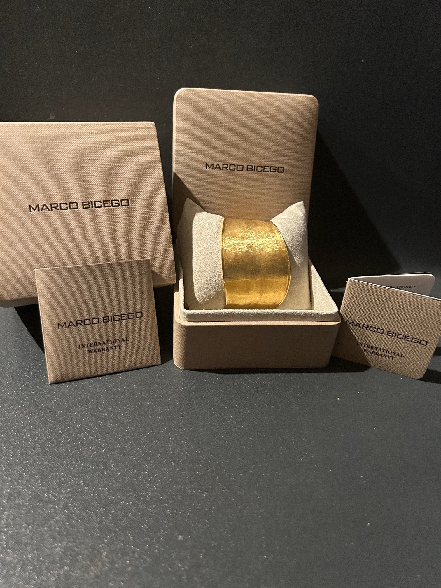 A Marco Bicego 18K gold cuff bracelet displayed in an open beige Marco Bicego box, with matching branded packaging and warranty cards arranged around it on a dark surface.