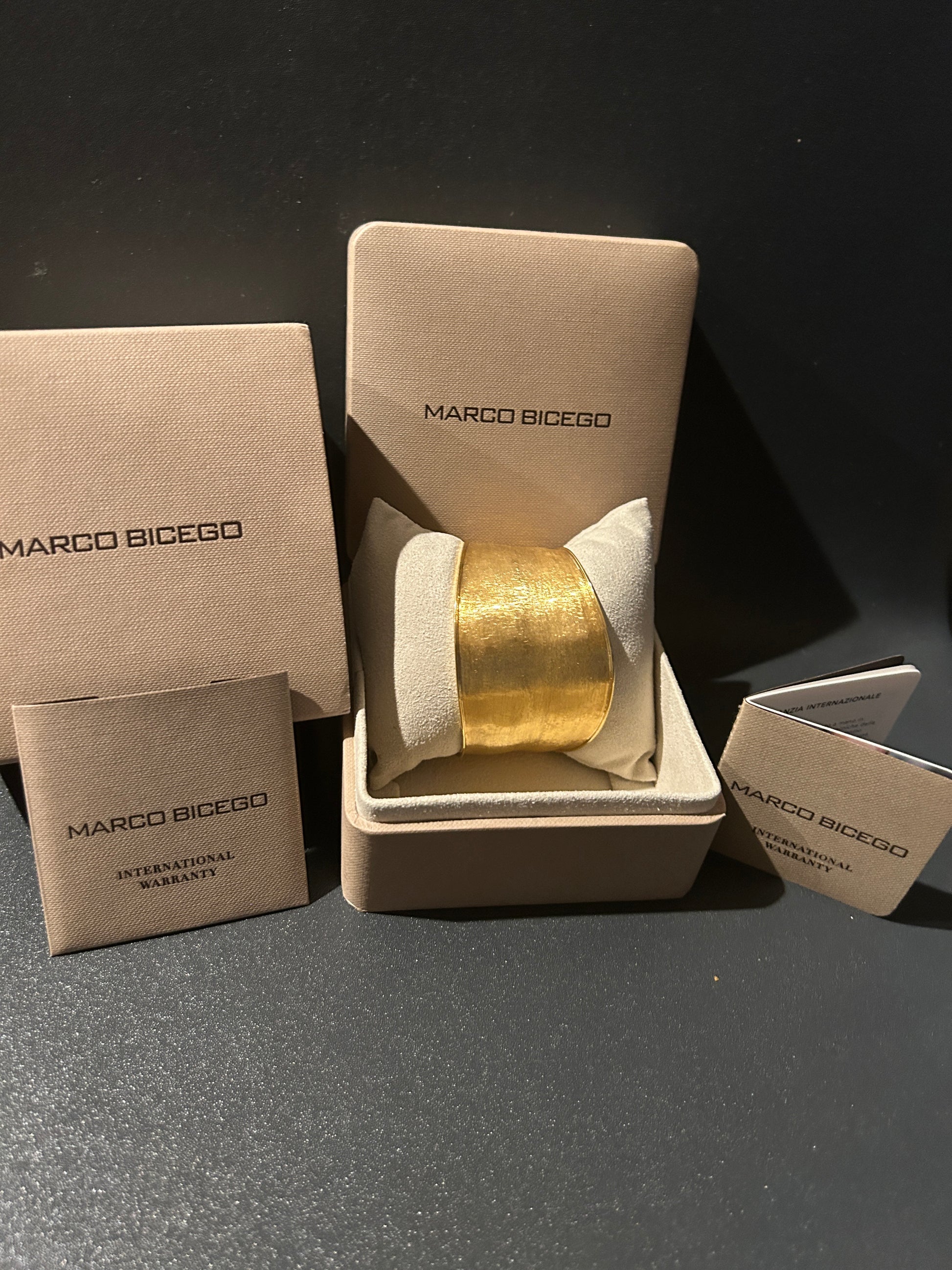 Marco Bicego 18K gold bracelet in its original beige presentation box, alongside its branded packaging and warranty card on a dark gray surface.