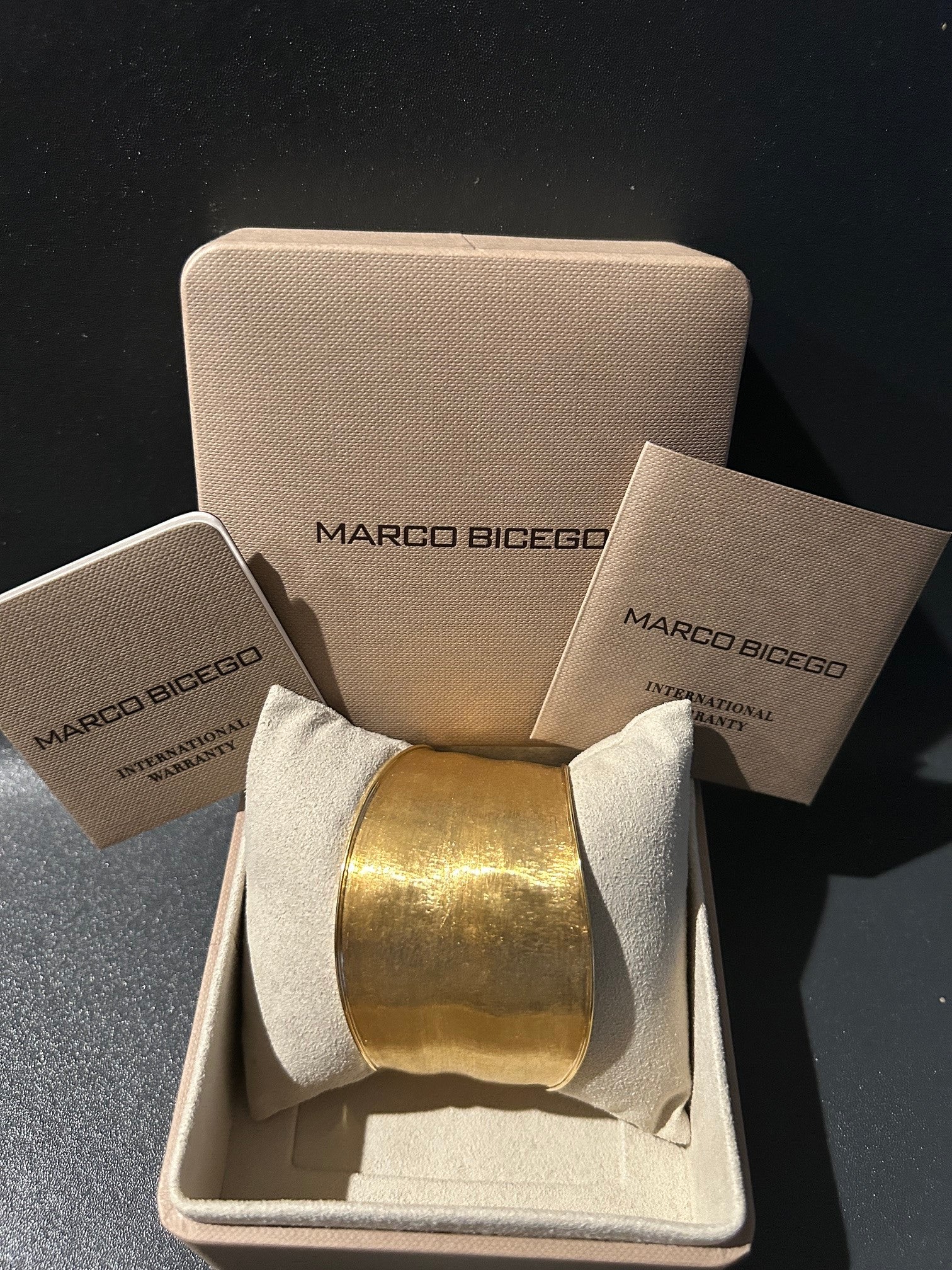 A Marco Bicego 18K gold cuff bracelet displayed inside an open beige jewelry box with branded inserts and a matching box lid behind it. The bracelet is centered on a cream pillow, with visible Marco Bicego branding on the packaging.