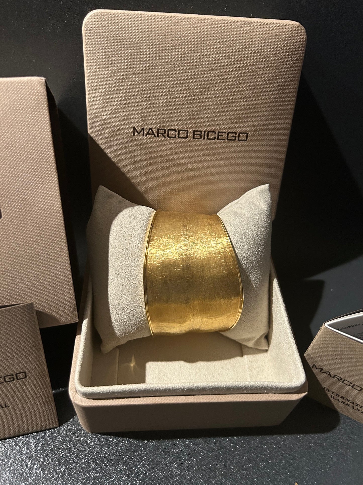 A Marco Bicego 18K gold cuff bracelet displayed inside an open beige Marcio Bicego jewelry box on a dark surface.