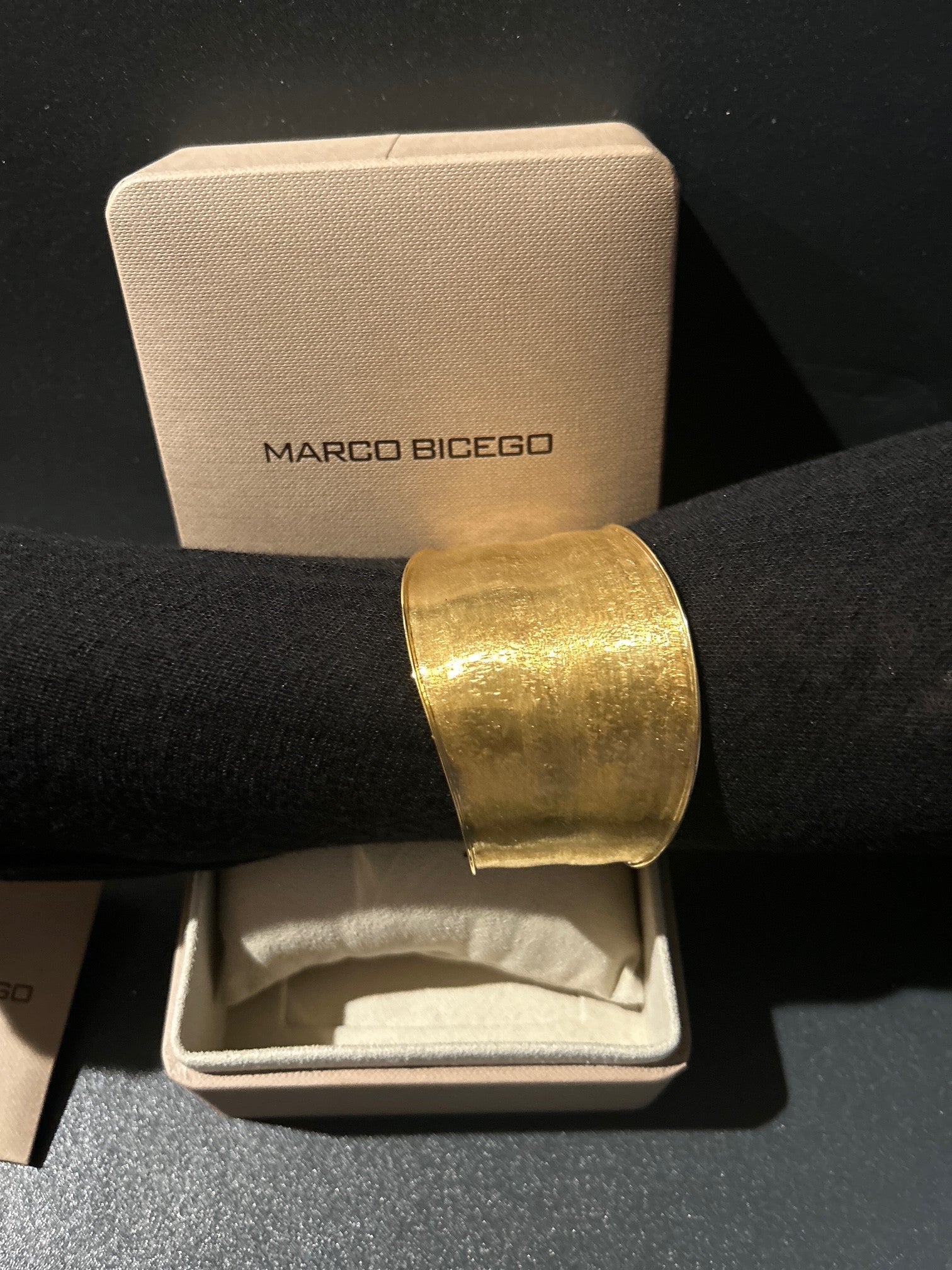 A Marco Bicego 18K gold cuff bracelet displayed on a black velvet wrist form, positioned in front of its beige Marcio Bicego jewelry box.