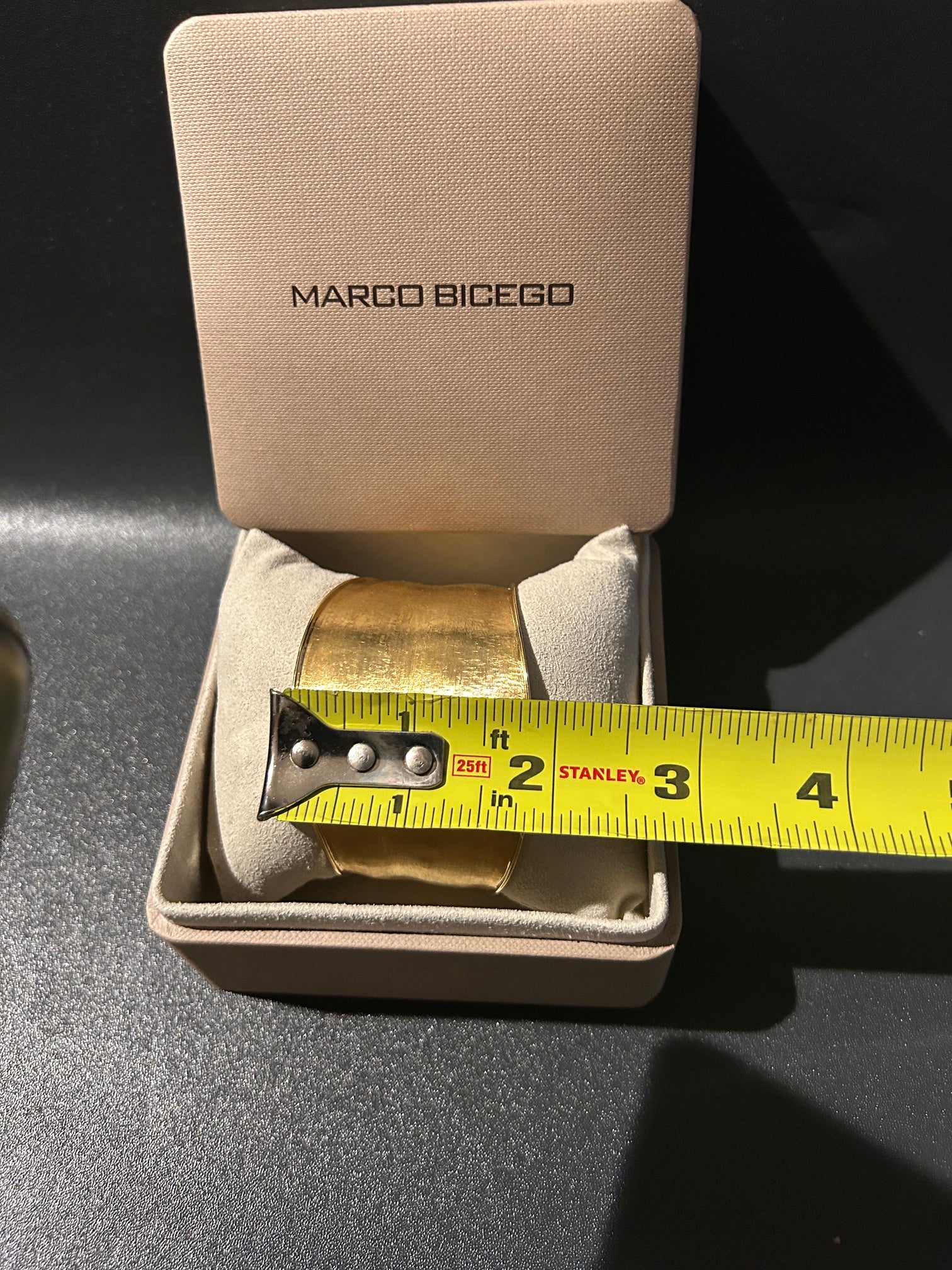 A Marco Bicego 18K gold cuff bracelet displayed inside its beige branded presentation box, with a yellow Stanley tape measure showing the width of the bracelet.