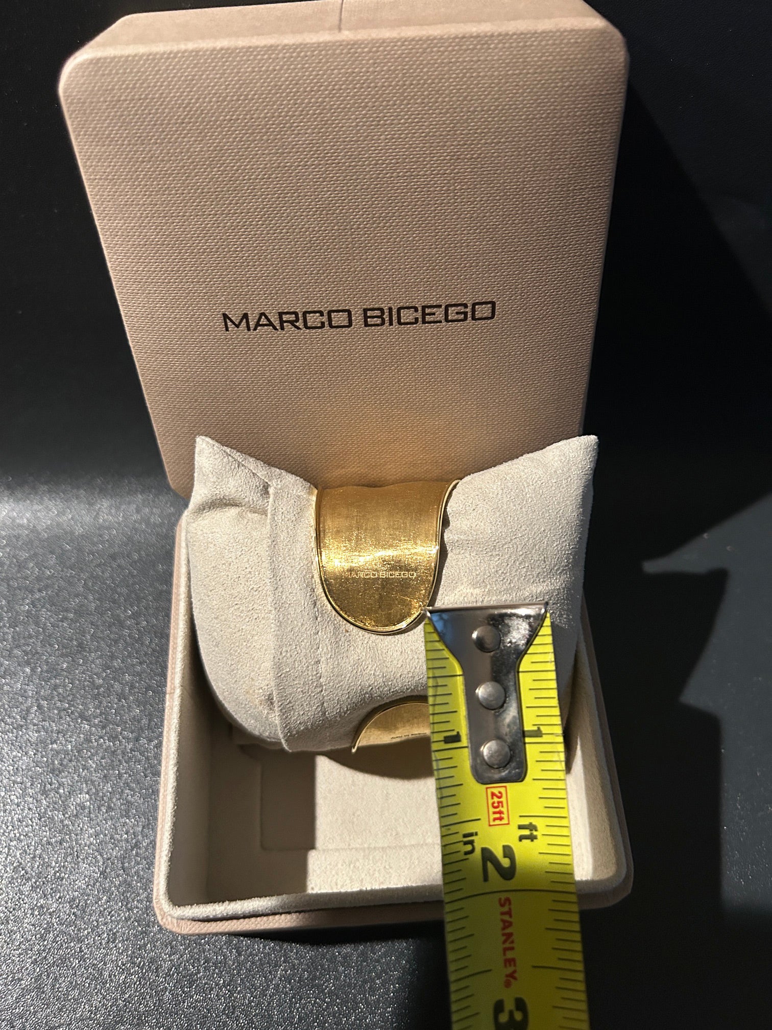 A Marco Bicego 18K gold cuff bracelet displayed inside its beige branded presentation box, with a yellow Stanley tape measure showing the width of the bracelet.