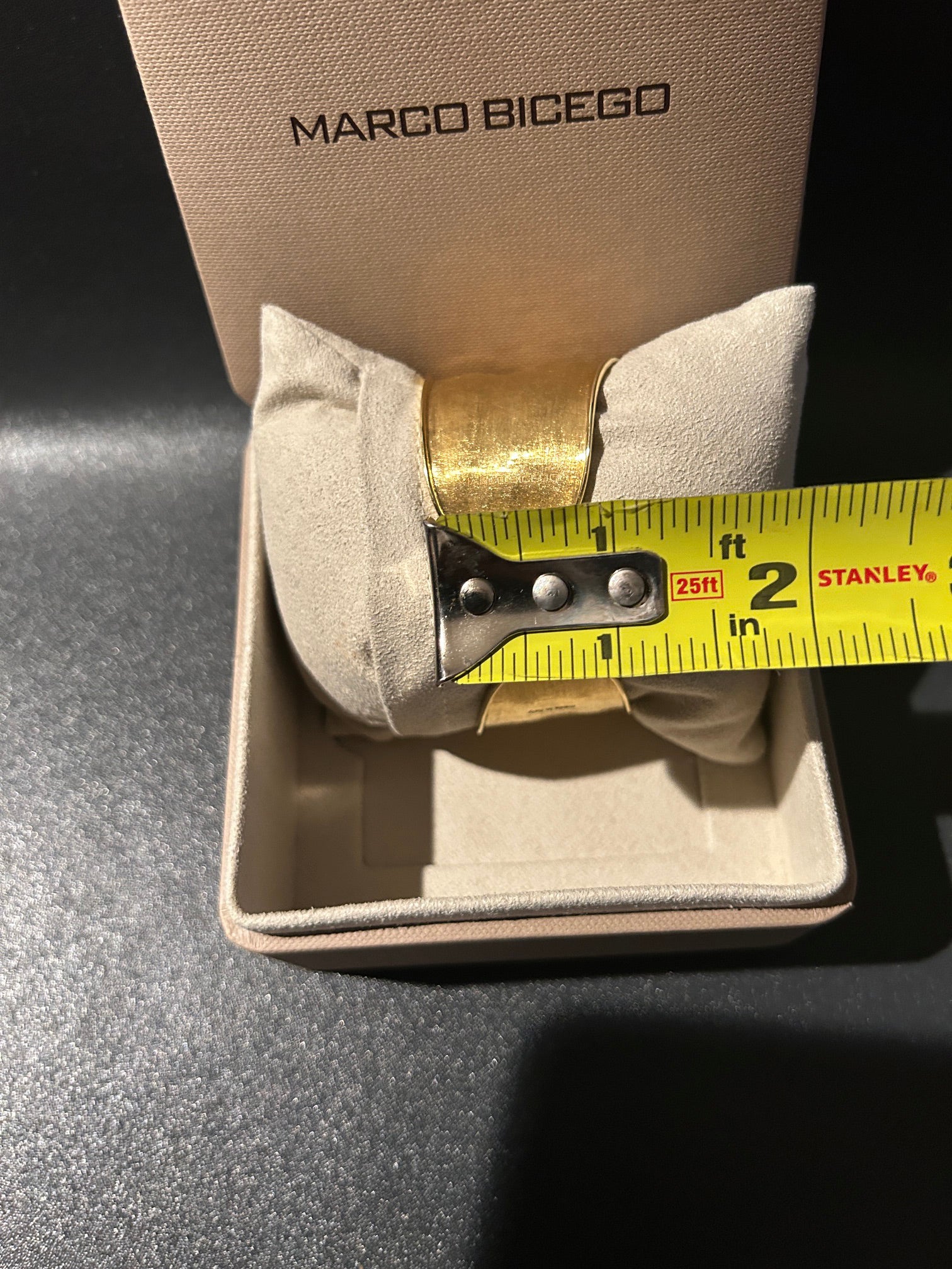 A Marco Bicego 18K gold cuff bracelet displayed inside its beige presentation box, resting on a beige cushion. A yellow Stanley tape measure is held across the top of the cushion to show the bracelet's width.